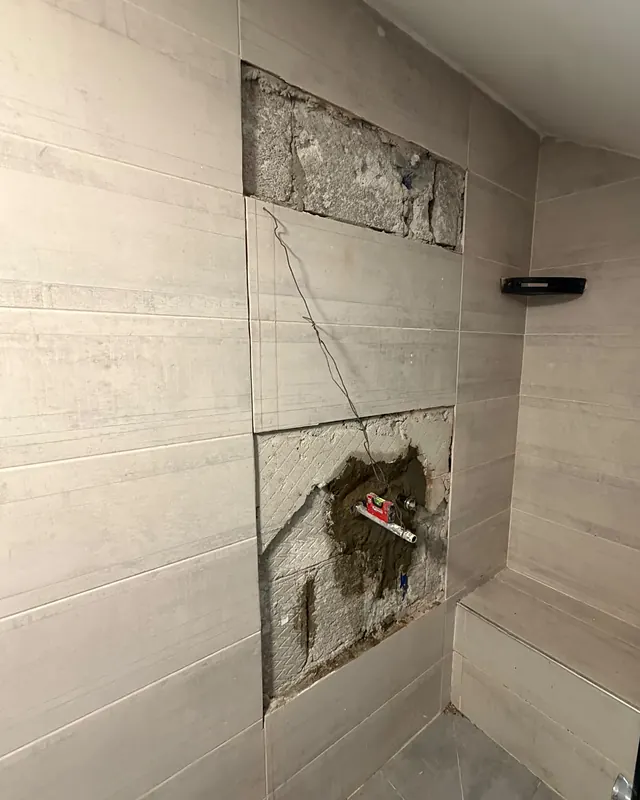 קיר מקלחת מחופה אריחים שבו הוסרו רק שני אריחים, זה מעל זה. בפתח התחתון נראים הצינור ופלס אדום, ושאר הקיר שלם